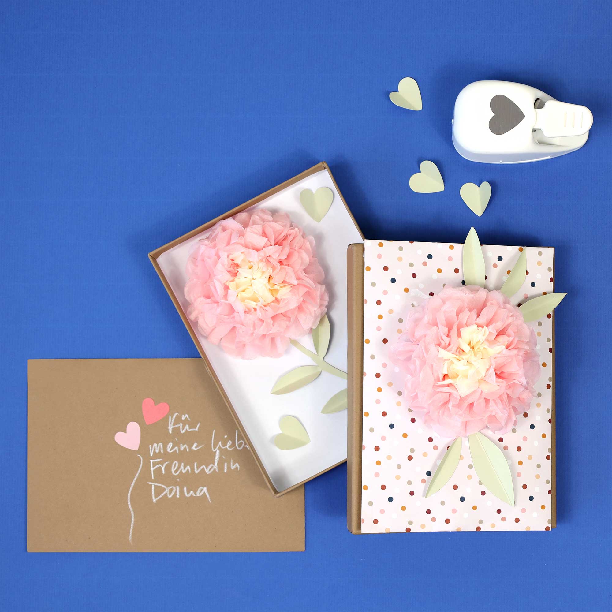 Une fleur en papier rose décore un paquet cadeau à pois sur fond bleu. À côté : une carte brune avec des cœurs et un texte en allemand, le Stanzer S Herz d'Artoz Papier AG, des cœurs en papier et du matériel de bricolage pour la fabrication de cartes.
