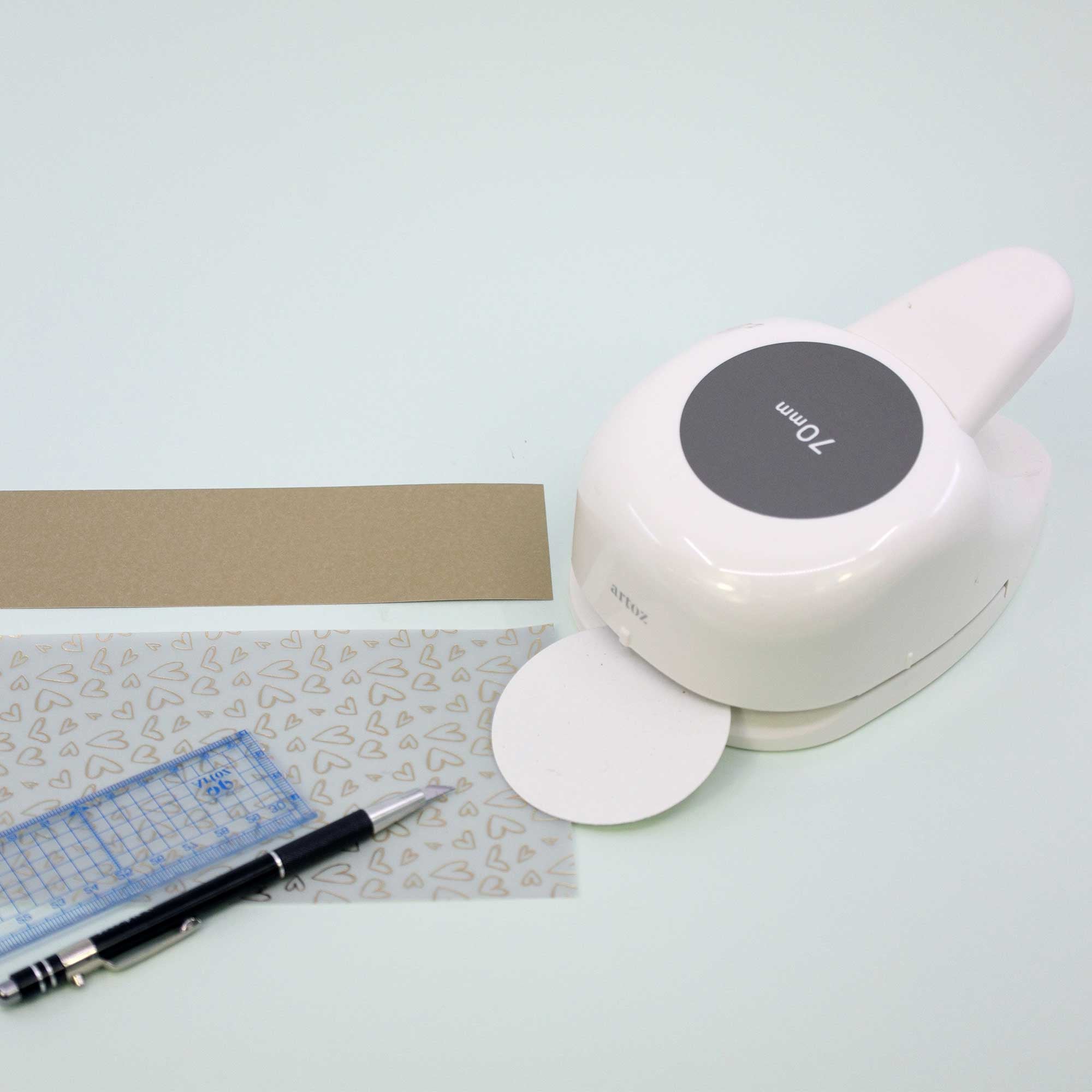 L'Artoz Papier AG Stanzer L Kreis (70 mm) est placé à côté d'une bande de papier brun, d'une règle claire, d'un stylo noir et d'une feuille avec des formes grises - des fournitures idéales pour les projets Basteln sur une surface blanche.