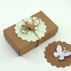 Une petite boîte cadeau marron avec de la ficelle, décorée avec du scrapbooking et une étiquette verte festonnée réalisée avec le Stanzer L Wellen-Kreis d'Artoz Papier AG, présente des motifs floraux et un lapin découpé. À côté se trouve une carte pour Karten basteln avec un papillon découpé.