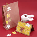 Un sac cadeau marron et une carte de vœux jaune présentent des feuilles découpées avec le Stanzer M Ahornblatt d'Artoz Papier AG, idéal pour les travaux manuels d'automne. Une ficelle rouge et jaune agrémente l'ensemble, présenté sur un fond rouge vif.