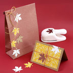 Un sac cadeau marron et une carte de vœux jaune présentent des feuilles découpées avec le Stanzer M Ahornblatt d'Artoz Papier AG, idéal pour les travaux manuels d'automne. Une ficelle rouge et jaune agrémente l'ensemble, présenté sur un fond rouge vif.