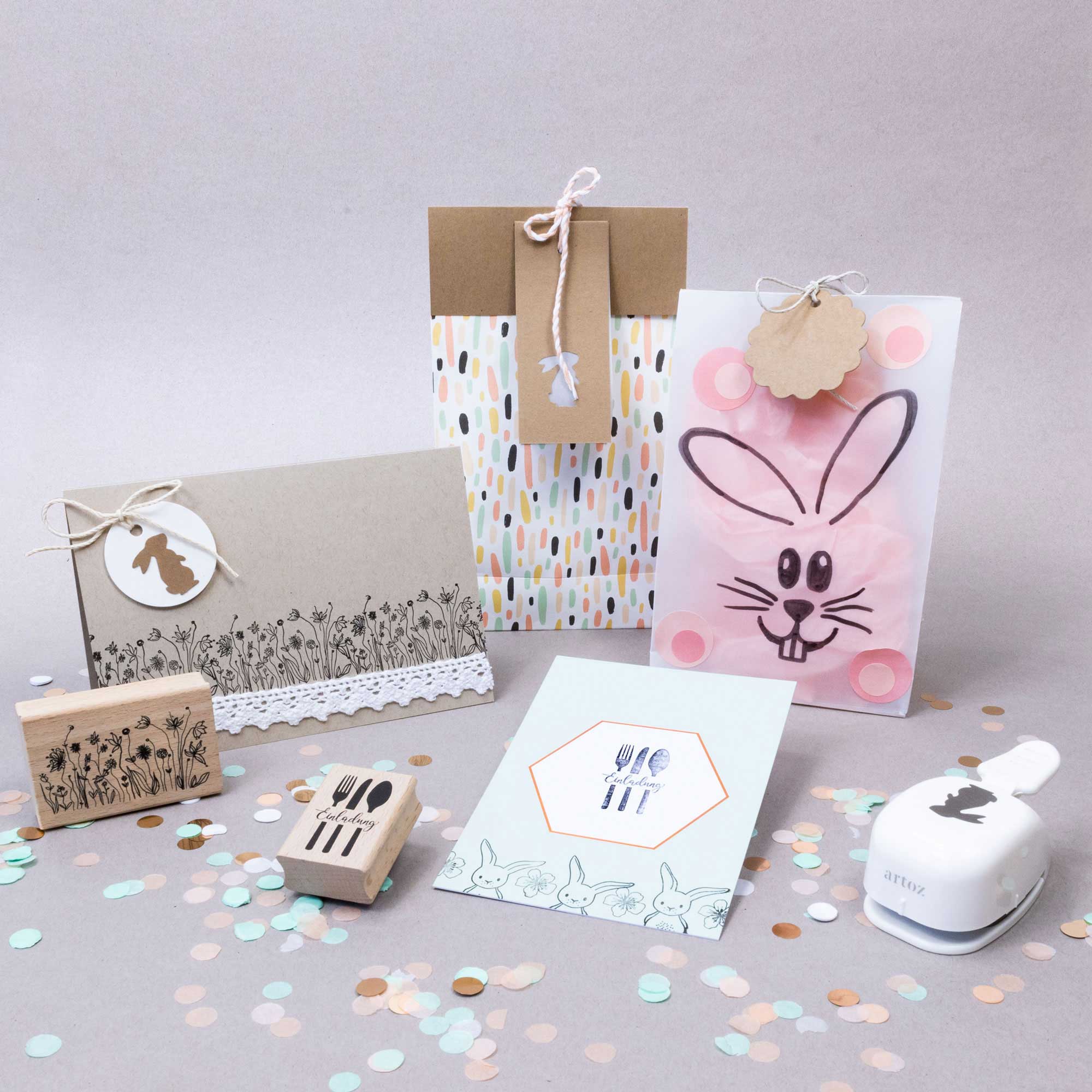 Des cartes de Pâques faites à la main, des sacs cadeaux et des étiquettes avec des motifs, des fleurs et des découpes de Hase sont présentés au milieu de confettis pastel. Des Bastelwerkzeug tels que des tampons en caoutchouc et l'Artoz Papier AG Stanzer S Hase sont également exposés sur une surface claire.