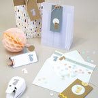 Des sacs cadeaux, des cartes et des enveloppes avec des découpes Hase Motiv et des confettis sont présentés sur un fond beige avec une boule rose en nid d'abeille, l'outil de perforation Artoz Papier AG Stanzer S Hase et des confettis de couleur pastel.