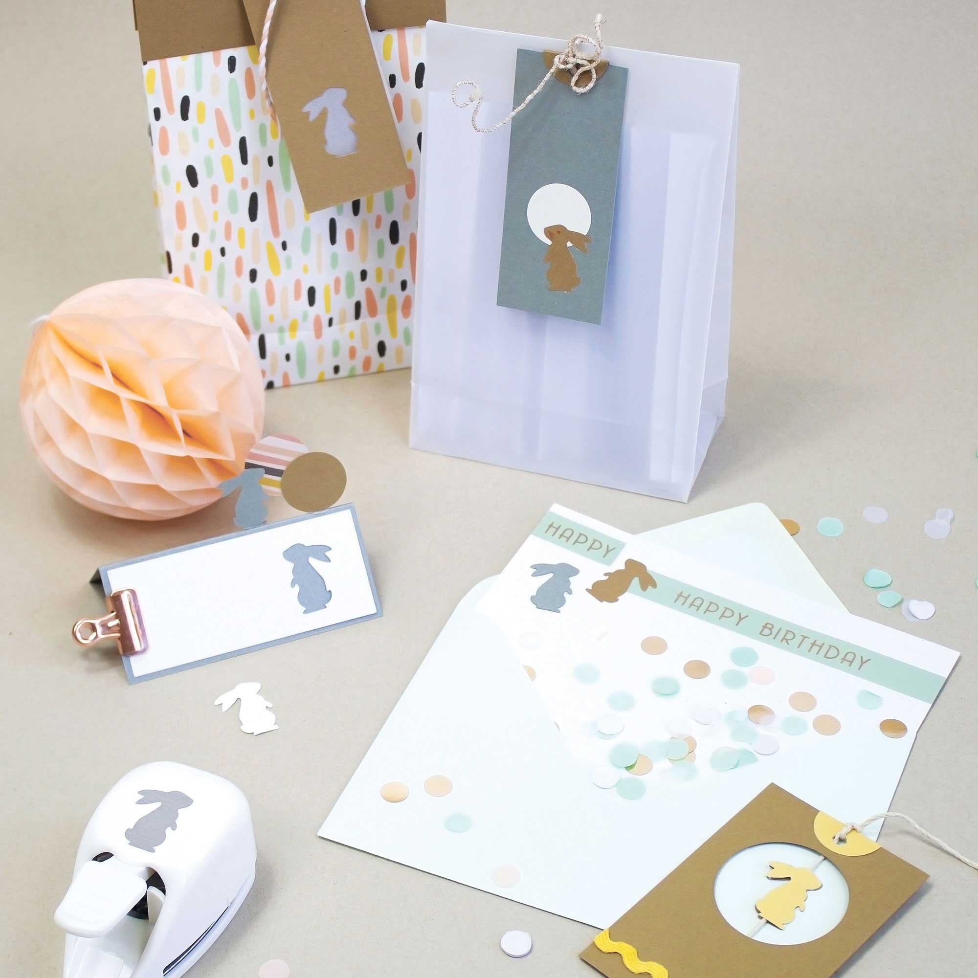 Des sacs cadeaux, des cartes et des enveloppes avec des découpes Hase Motiv et des confettis sont présentés sur un fond beige avec une boule rose en nid d'abeille, l'outil de perforation Artoz Papier AG Stanzer S Hase et des confettis de couleur pastel.