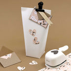 Un sac cadeau blanc avec des découpes de cœurs floraux, une carte "Pour toi", l'outil de perforation Artoz Papier AG Stanzer S Herz asymétrique, ainsi que des cœurs en papier et des enveloppes - idéal pour les projets de scrapbooking ou de design sur fond marron.