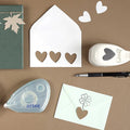Créez une charmante scène Bastel-Design avec le Stanzer S Herz asymétrique d'Artoz Papier AG, une enveloppe blanche avec des découpes en forme de cœur, un stylo, un dévidoir de ruban adhésif, des cœurs en papier, une enveloppe verte avec des découpes en forme de cœur et un carnet vert avec des découpes en forme de feuille.