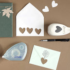 Créez une charmante scène Bastel-Design avec le Stanzer S Herz asymétrique d'Artoz Papier AG, une enveloppe blanche avec des découpes en forme de cœur, un stylo, un dévidoir de ruban adhésif, des cœurs en papier, une enveloppe verte avec des découpes en forme de cœur et un carnet vert avec des découpes en forme de feuille.