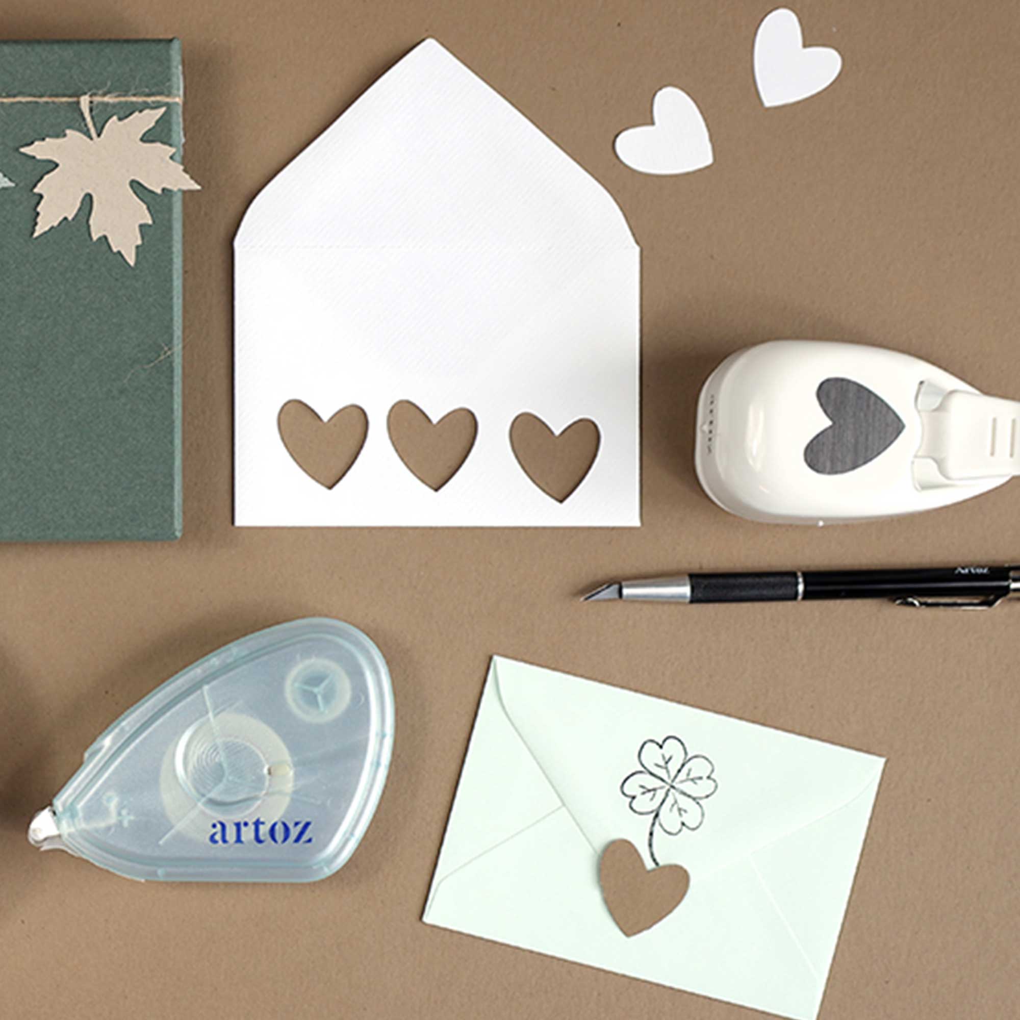 Créez une charmante scène Bastel-Design avec le Stanzer S Herz asymétrique d'Artoz Papier AG, une enveloppe blanche avec des découpes en forme de cœur, un stylo, un dévidoir de ruban adhésif, des cœurs en papier, une enveloppe verte avec des découpes en forme de cœur et un carnet vert avec des découpes en forme de feuille.