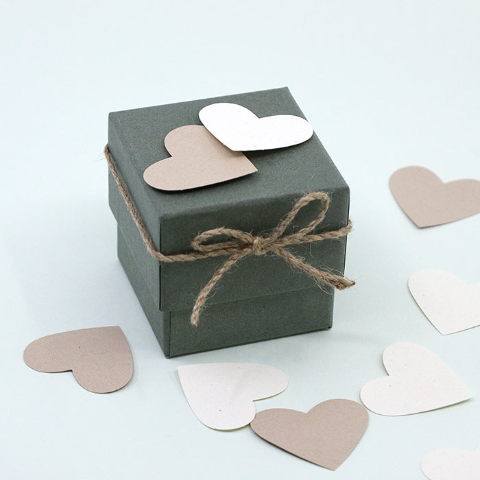 Une petite boîte cadeau vert foncé, attachée avec de la ficelle, est décorée de cœurs en papier réalisés à l'aide de l'Artoz Papier AG Stanzer M Herz. Des découpes de cœurs beiges et blancs sont disséminées un peu partout, idéales pour les Karten basteln.