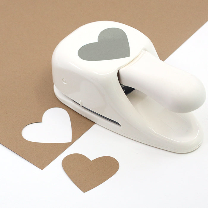 Un Stanzer M Herz blanc d'Artoz Papier AG repose sur du papier brun, présentant une découpe en forme de cœur et le cœur en papier à côté - idéal pour la fabrication de cartes (Karten basteln) et les projets créatifs.