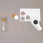 Un pot en verre transparent avec un couvercle en liège, du papier à motifs, du rickrack rose, des cercles festonnés, une étiquette perforée et des outils Basteln comme le Artoz Papier AG Stanzer M Kreis sont disposés sur une surface grise.