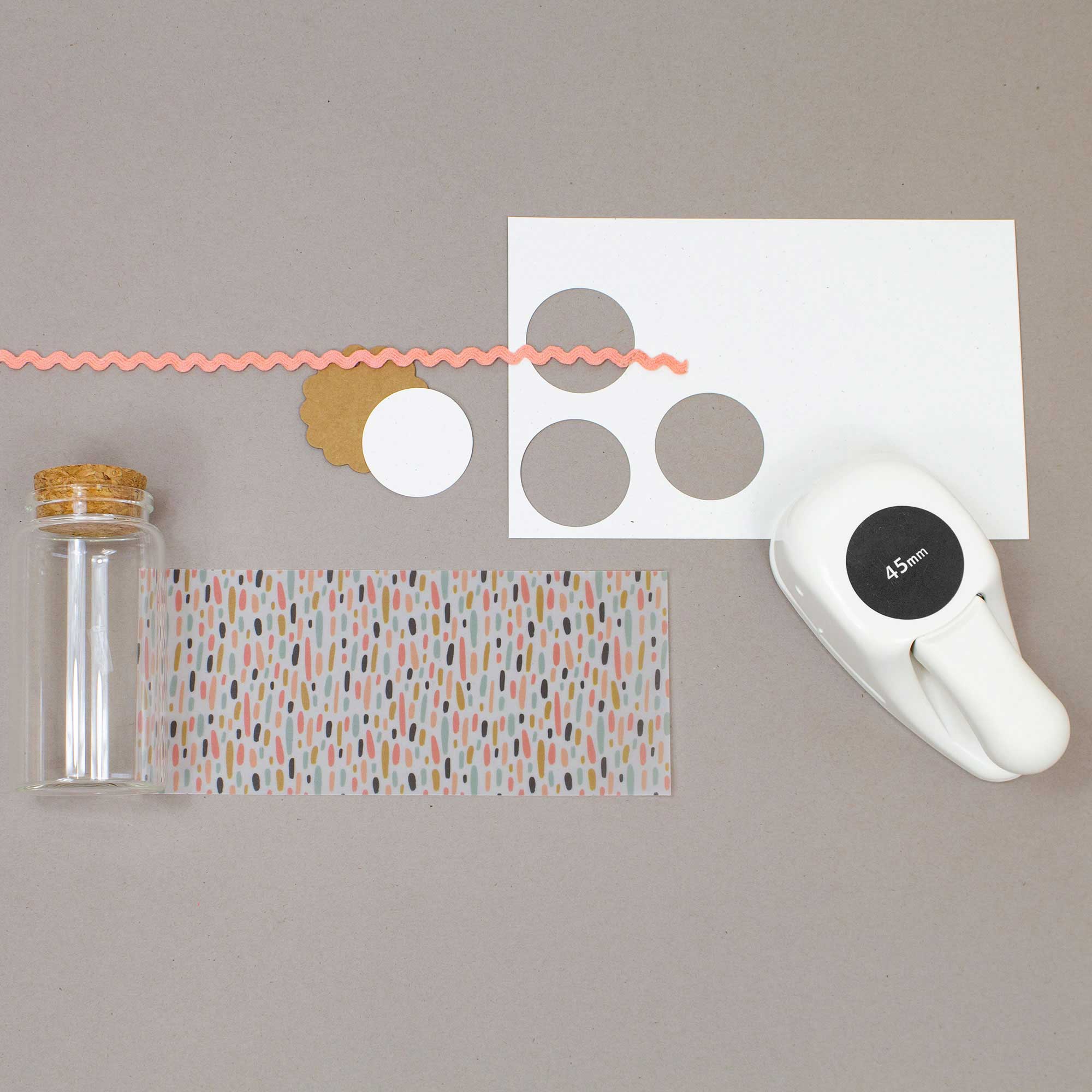 Un pot en verre transparent avec un couvercle en liège, du papier à motifs, du rickrack rose, des cercles festonnés, une étiquette perforée et des outils Basteln comme le Artoz Papier AG Stanzer M Kreis sont disposés sur une surface grise.