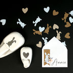 La perforatrice Artoz Papier AG Stanzer M Osterhase, en forme de lapin, est posée sur une surface noire avec des découpes éparses. À proximité se trouvent des perforatrices en forme de cœur, une enveloppe avec une carte "Annie" et le Bastelpapier Osterdeko.
