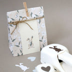 Un sac cadeau fleuri avec un nœud en ficelle et un lapin découpé se trouve à côté de Bastelpapier Osterdeko, de papiers découpés en forme de cœur et de lapin, et de l'Artoz Papier AG Stanzer M Osterhase sur un fond neutre.