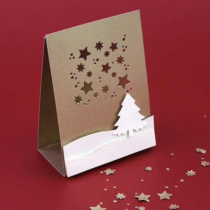 Le Fliegender Stanzer Sterne d'Artoz Papier AG est un sac lumineux or et blanc pour Basteln, représentant un sapin de Noël découpé sur une colline enneigée avec des étoiles, sur fond rouge avec des confettis d'étoiles.