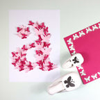Sur une feuille blanche figure un grand chiffre 2 composé de papillons rouges et roses superposés. À proximité se trouvent Artoz Papier AG Stanzer S Schmetterling et un carré magenta avec des découpes de papillons - idéal Bastelzubehör for Karten gestalten.