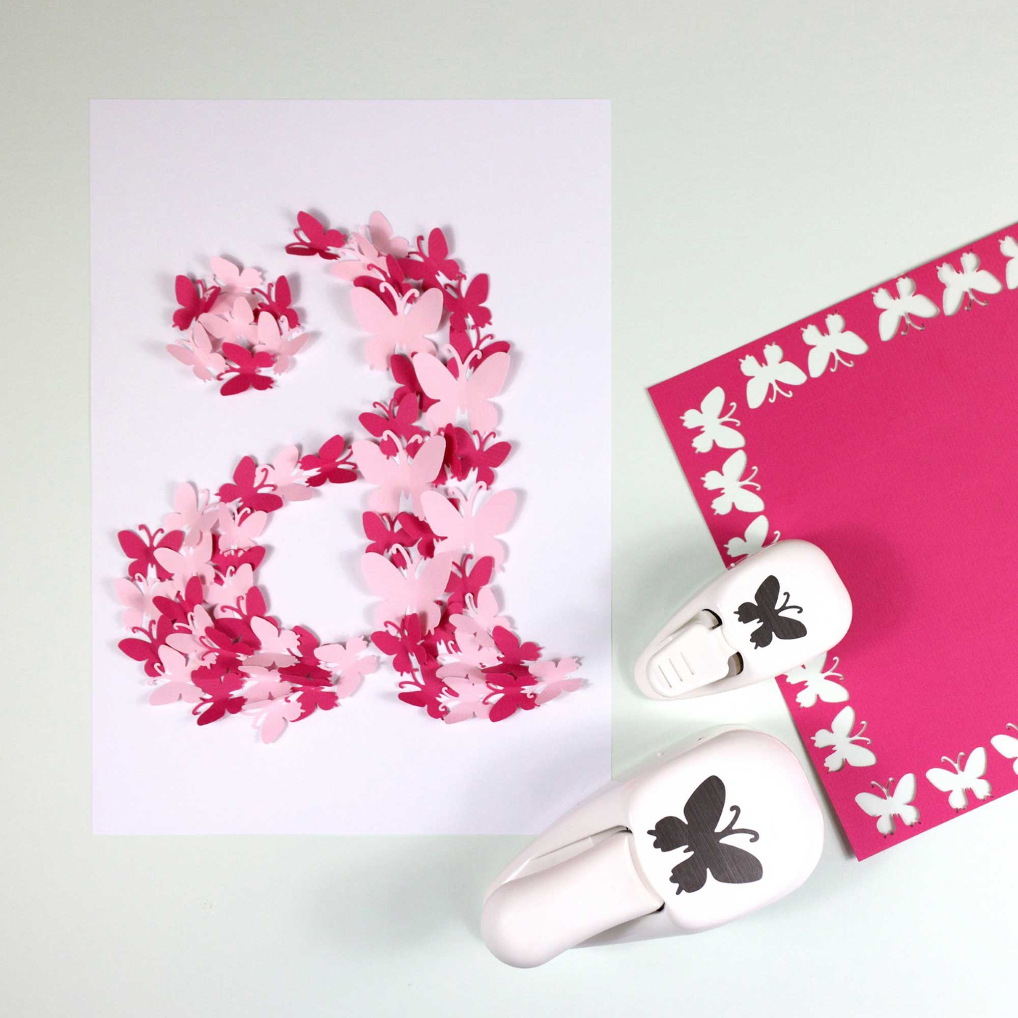Sur une feuille blanche figure un grand chiffre 2 composé de papillons rouges et roses superposés. À proximité se trouvent Artoz Papier AG Stanzer S Schmetterling et un carré magenta avec des découpes de papillons - idéal Bastelzubehör for Karten gestalten.