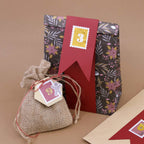 Eine Geschenktüte aus dem 6er-Set „Papiertüten Winter Design“ der Artoz Papier AG in verschiedenen Farben, gefertigt aus FSC-zertifiziertem Papier und mit einem Blumenmuster sowie einem roten Anhänger mit der Aufschrift „3“, befindet sich hinter einem kleinen, mit einer Schnur verschnürten Jutesack mit der Aufschrift „9“ – alles vor einem neutralen braunen Hintergrund.