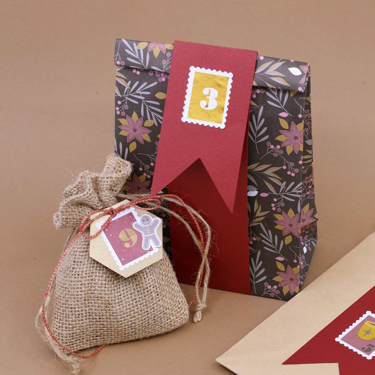 Eine Geschenktüte aus dem 6er-Set „Papiertüten Winter Design“ der Artoz Papier AG in verschiedenen Farben, gefertigt aus FSC-zertifiziertem Papier und mit einem Blumenmuster sowie einem roten Anhänger mit der Aufschrift „3“, befindet sich hinter einem kleinen, mit einer Schnur verschnürten Jutesack mit der Aufschrift „9“ – alles vor einem neutralen braunen Hintergrund.