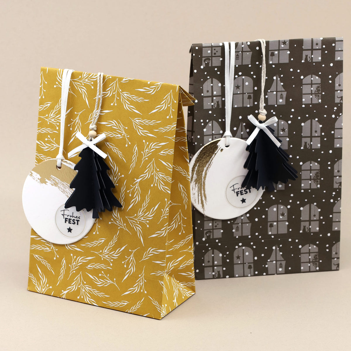 Neben einander standen zwei festliche Geschenktüten aus dem 6er-Set „Papiertüten Winter Design“ der Artoz Papier AG in verschiedenen Farben. Die eine hat einen senfgelben Hintergrund mit einem botanischen Motiv, während die andere braun ist und mit einem Haus- und Schneeflockenmuster verziert ist. Beide Tüten sind mit runden Anhängern versehen und mit kleinen schwarzen Baumschmuckstücken geschmückt, die mit einer Schnur befestigt sind.
