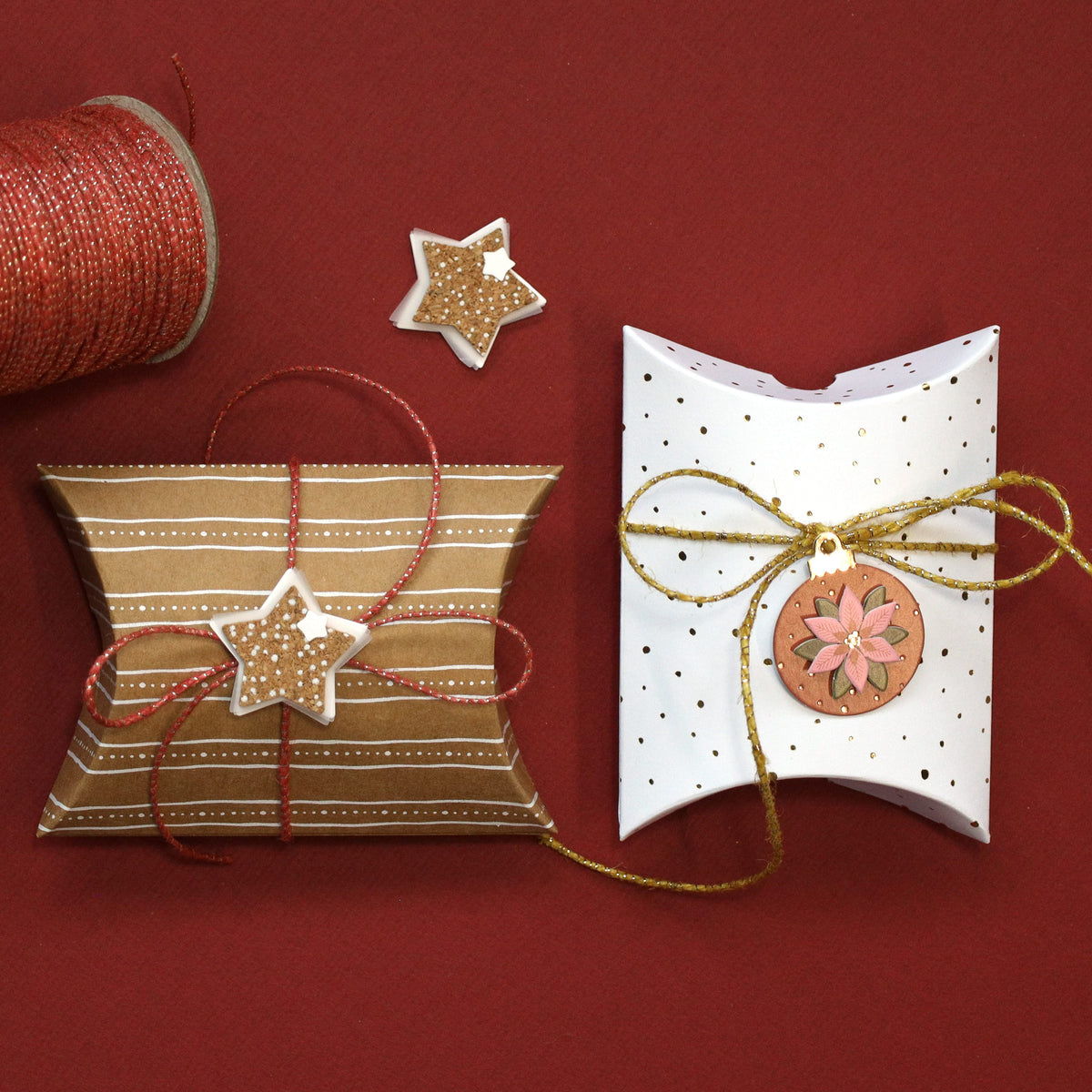 Deux petites boîtes-cadeaux d'Artoz Papier AG, les « Boîtes pliantes de Noël aux étoiles » – l'une à rayures dorées et l'autre blanche à pois dorés – sont ornées d'étiquettes festives représentant des étoiles et des fleurs. Une bobine de ficelle rouge et dorée et un petit ornement en forme d'étoile reposent sur un fond rouge vif – l'idéal pour créer d'élégants emballages cadeaux pour toutes les occasions.