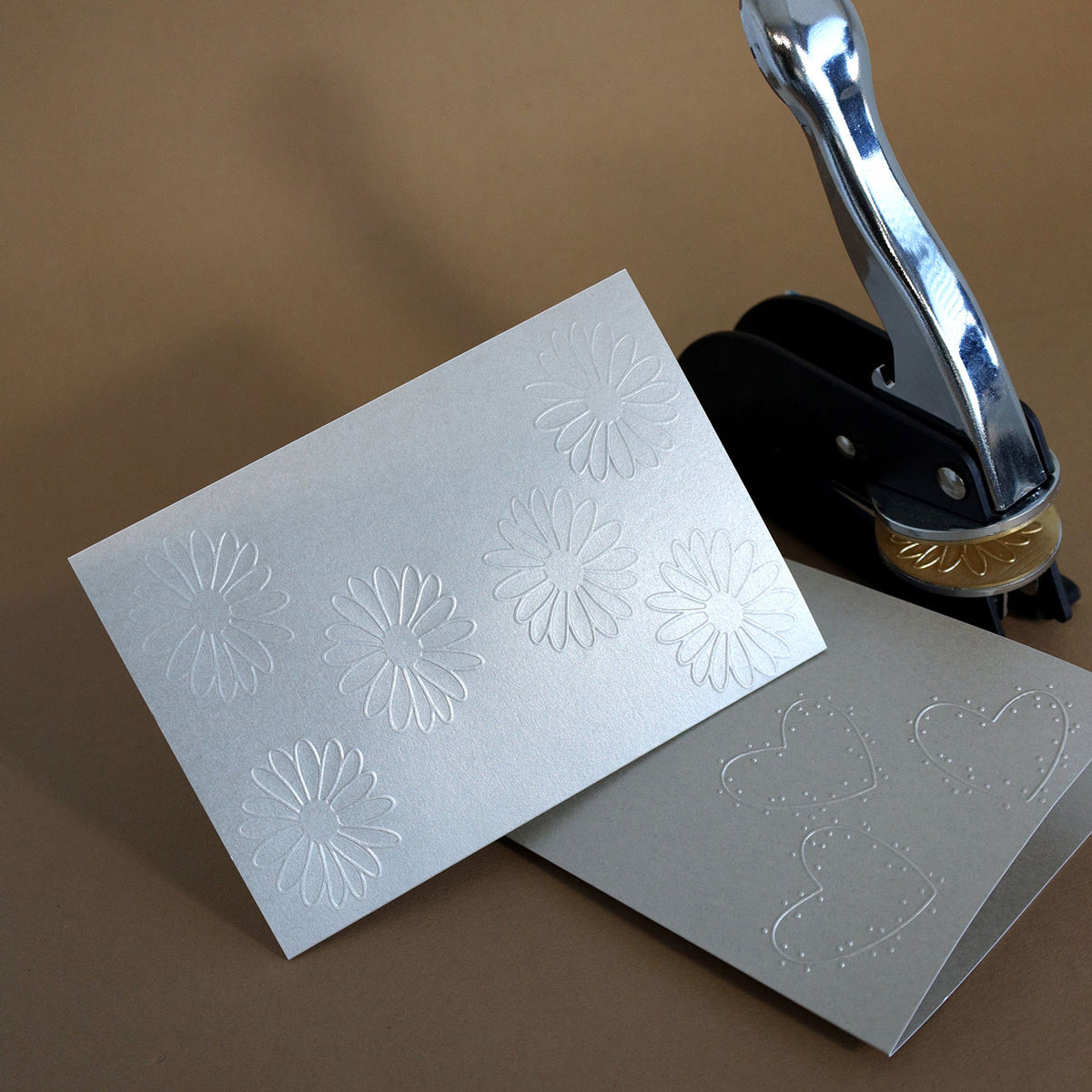 On peut voir deux cartes argentées ornées de motifs floraux et de cœurs en relief. À côté d'elles se trouve l'outil d'estampage « Prägestempel Ersatz Sujet Blume » de la société Artoz Papier AG, reconnaissable à son sceau doré, le tout disposé sur un fond marron.