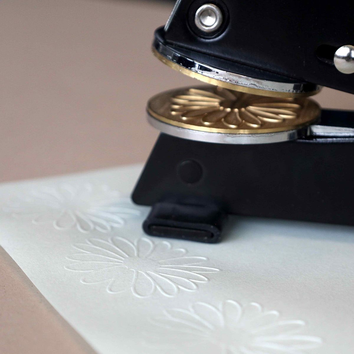 Gros plan sur un tampon à gaufrer noir « Prägestempel Ersatz Sujet Blume » de la marque Artoz Papier AG, en train d'imprimer un motif floral sur du papier blanc et de créer des motifs floraux en relief.