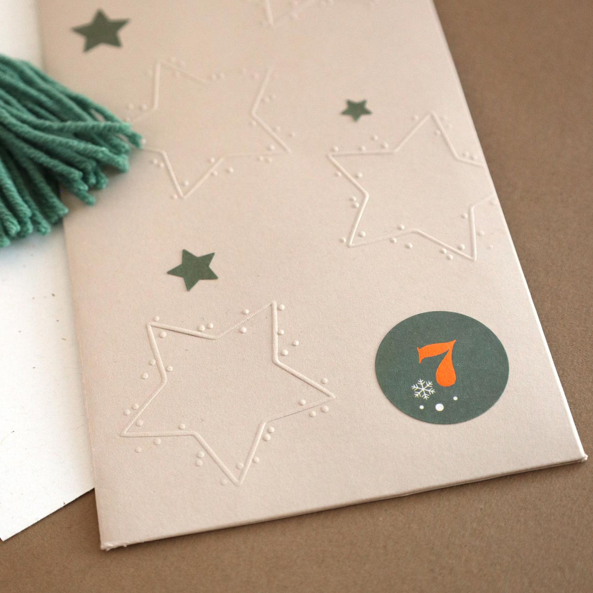 Une carte beige est ornée d'étoiles en relief réalisées à l'aide du tampon à gaufrer « Motiv Stern » d'Artoz Papier AG, rehaussées d'autocollants verts en forme d'étoile et d'un autocollant rond représentant un chiffre 7 orange agrémenté d'un petit flocon de neige blanc. Un pompon en fil vert apporte une touche originale, idéale pour vos projets de bricolage ou pour vous inspirer dans votre prochain projet créatif.