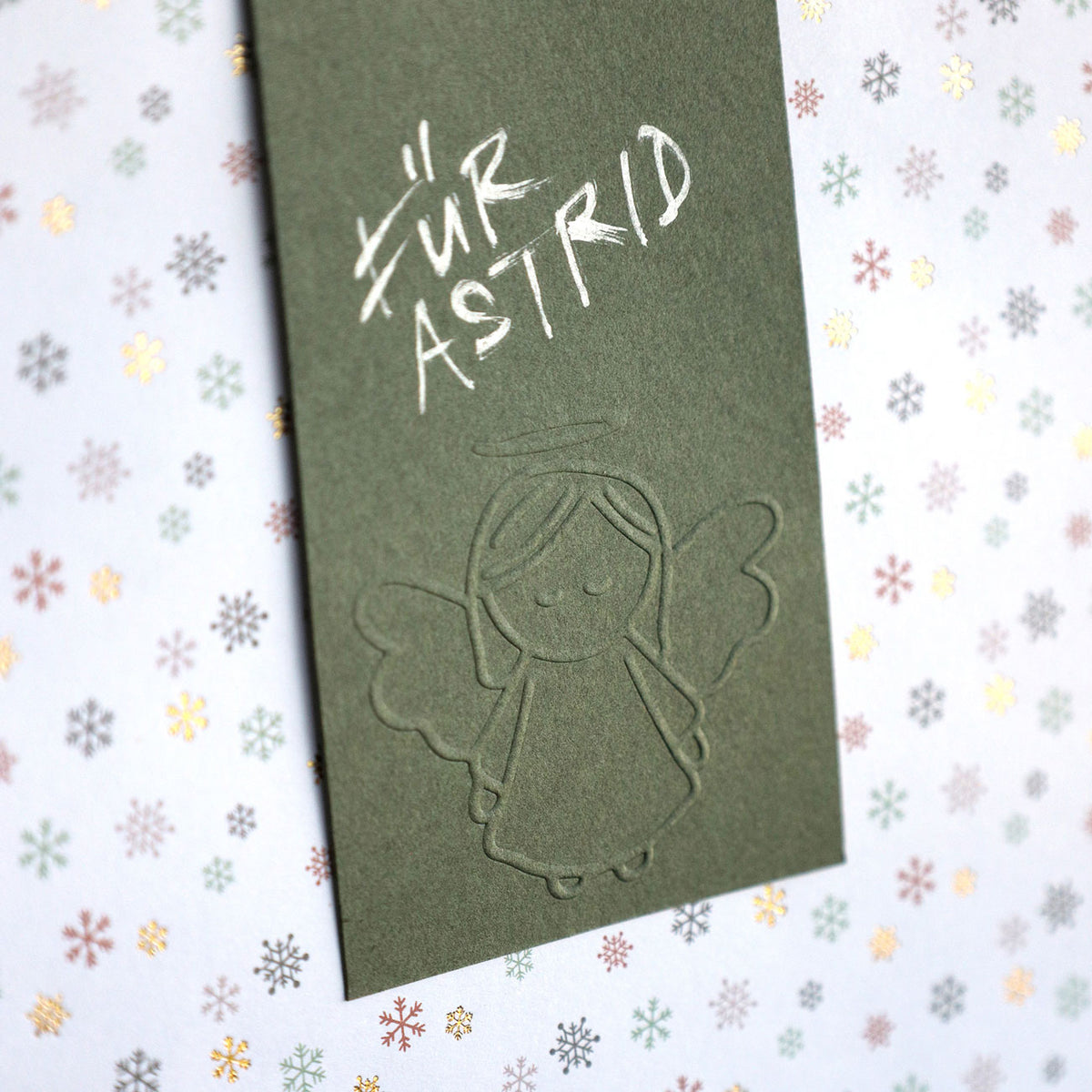 Une carte verte ornée d'un ange en relief réalisé à l'aide du tampon de gaufrage « Ersatz Sujet Engel » d'Artoz Papier AG, avec une inscription manuscrite en blanc « Für Astrid », posée sur du papier à motifs de flocons de neige : un charmant exemple de projet de bricolage utilisant des cartes gaufrées pour apporter une touche personnelle.