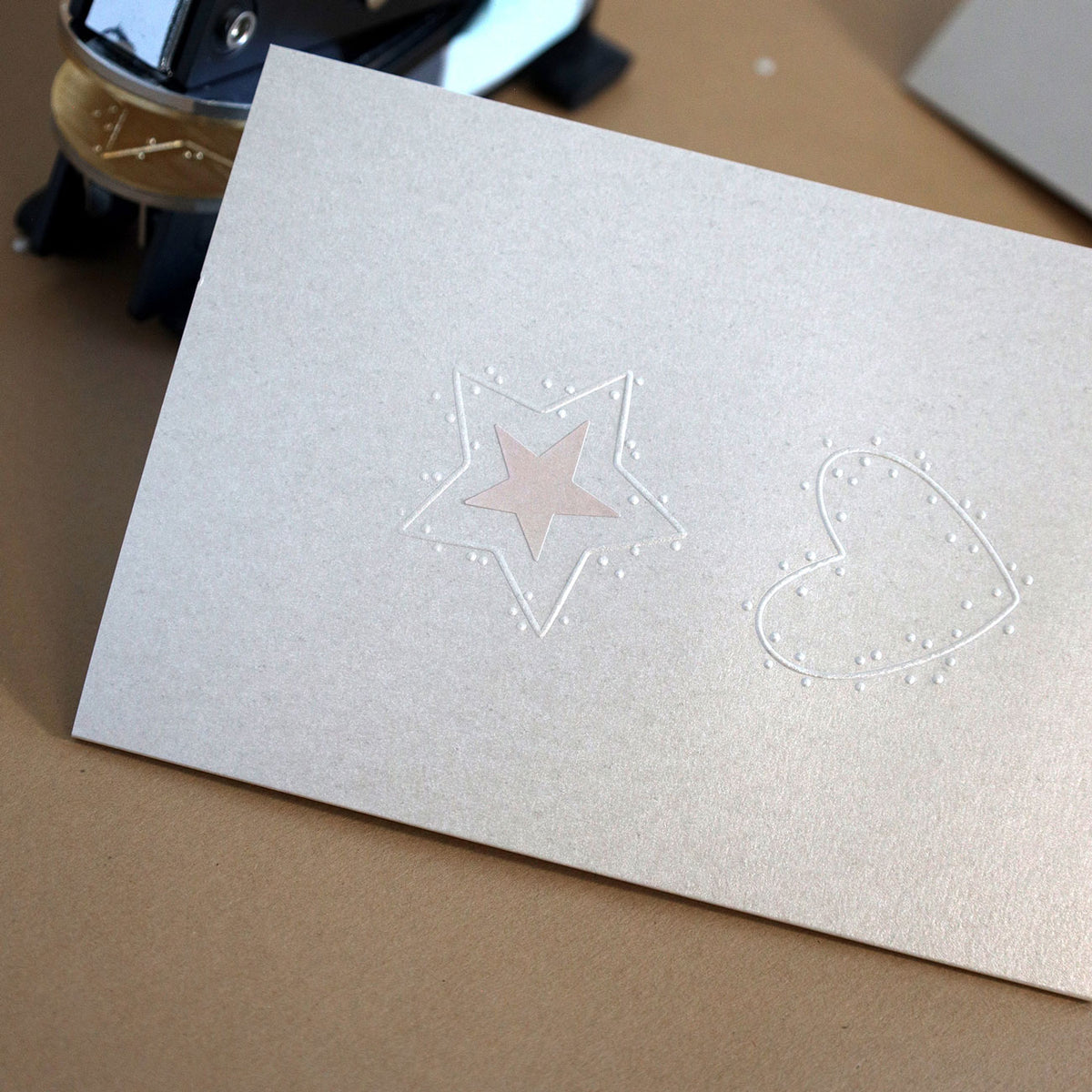 Une carte beige ornée de motifs en relief représentant des étoiles et des cœurs — réalisés à l'aide du tampon à gaufrer « Motiv Herz » d'Artoz Papier AG — repose sur un fond marron. À l'arrière-plan, on aperçoit partiellement un dévidoir de ruban adhésif, qui vous donnera l'inspiration parfaite pour vos prochains projets de bricolage.