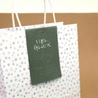 Un sac cadeau blanc, orné d'un délicat motif de petites fleurs et doté de poignées blanches, est agrémenté d'une étiquette verte sur laquelle on peut lire « Viel Glück » et d'un trèfle à quatre feuilles en relief, réalisé à l'aide du tampon de gaufrage « Prägestempel Ersatz Sujet Kleeblatt » de la marque Artoz Papier AG.