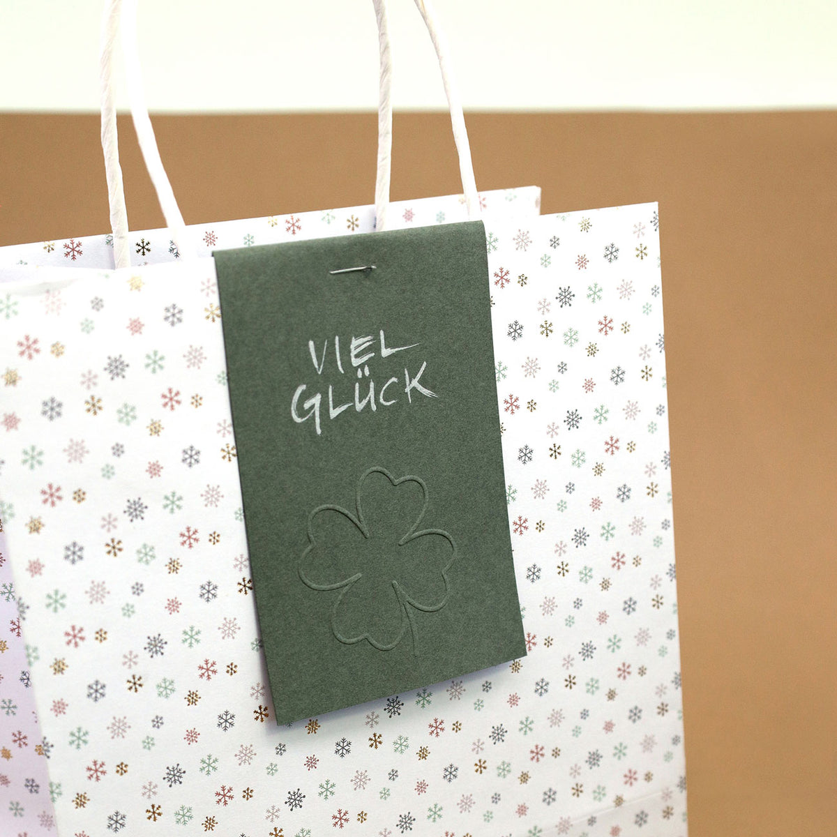 Un sac cadeau blanc, orné d'un délicat motif de petites fleurs et doté de poignées blanches, est agrémenté d'une étiquette verte sur laquelle on peut lire « Viel Glück » et d'un trèfle à quatre feuilles en relief, réalisé à l'aide du tampon de gaufrage « Prägestempel Ersatz Sujet Kleeblatt » de la marque Artoz Papier AG.