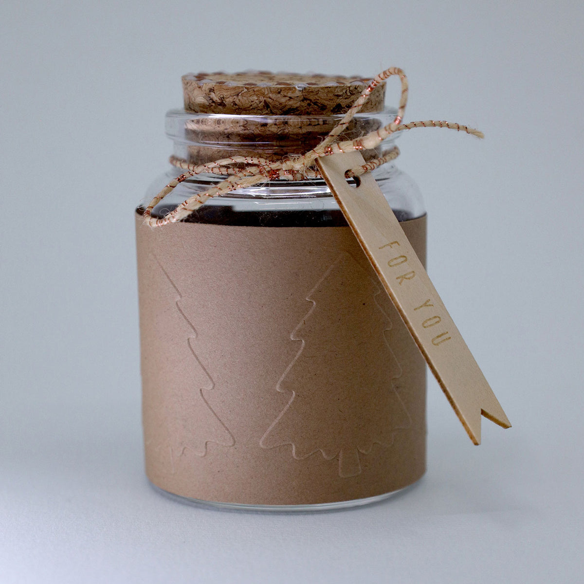 Un petit bocal en verre muni d'un couvercle en liège, enveloppé dans du papier kraft sur lequel est estampé le motif « Tannenbaum » à l'aide du tampon « Prägestempel Ersatz Sujet Tannenbaum » de la marque Artoz Papier AG, et noué avec de la ficelle. Une étiquette attachée à la ficelle porte l'inscription « FOR YOU ».