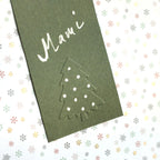 Une carte verte sur laquelle figure le mot « Mami » en blanc, avec un sapin de Noël en contour en dessous, réalisée à l'aide du tampon en relief « Prägestempel Ersatz Sujet Tannenbaum » de la marque Artoz Papier AG, posée sur du papier orné de petits flocons de neige aux couleurs pastel.