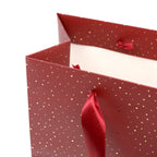 Gros plan sur le sac cadeau « Geschenk Tragtasche Pure M Weihnachtlich rot gold » d'Artoz Papier AG, un sac cadeau rouge orné de pois dorés, doté de poignées en ruban de satin rouge et d'un intérieur blanc — idéal pour un emballage cadeau élégant.