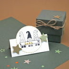 Un porte-nom orné de trois lutins illustrés issus de la série « Stempel Wichtel » d’Artoz Papier AG, personnalisé avec le prénom « Sara » et décoré de deux étoiles dorées, repose sur du papier vert issu de fournitures de loisirs créatifs haut de gamme. À l'arrière-plan, une boîte-cadeau verte fermée par un cordon et des confettis en forme d'étoiles parsemés ici et là contribuent à créer une ambiance festive. Pour encore plus de charme, d'autres touches décoratives, telles qu'un « tampon sapin » ou un «