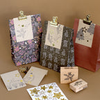 Drei verzierte Geschenktüten mit festlichen Motiven und Namensschildern liegen auf einer braunen Unterlage, umgeben von goldenen Klammern, Sternenaufklebern, dem Stempelset „Floral Winter“ von Artoz Papier AG, einem Stempelkissen und illustrierten Karten – alles für ein bezauberndes Bastelprojekt zur Weihnachtszeit.