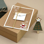 Une carte de Noël ornée de motifs représentant des souris et des cadeaux, accompagnée d'une enveloppe miniature, repose sur un coffret cadeau marron : une touche de choix pour les amateurs d'emballages cadeaux. À côté se trouve le « Stempel Geschenk klein » d'Artoz Papier AG, avec son petit sapin de Noël en papier vert surmonté d'une étoile : une scène charmante qui inspirera vos créations manuelles pour les fêtes.