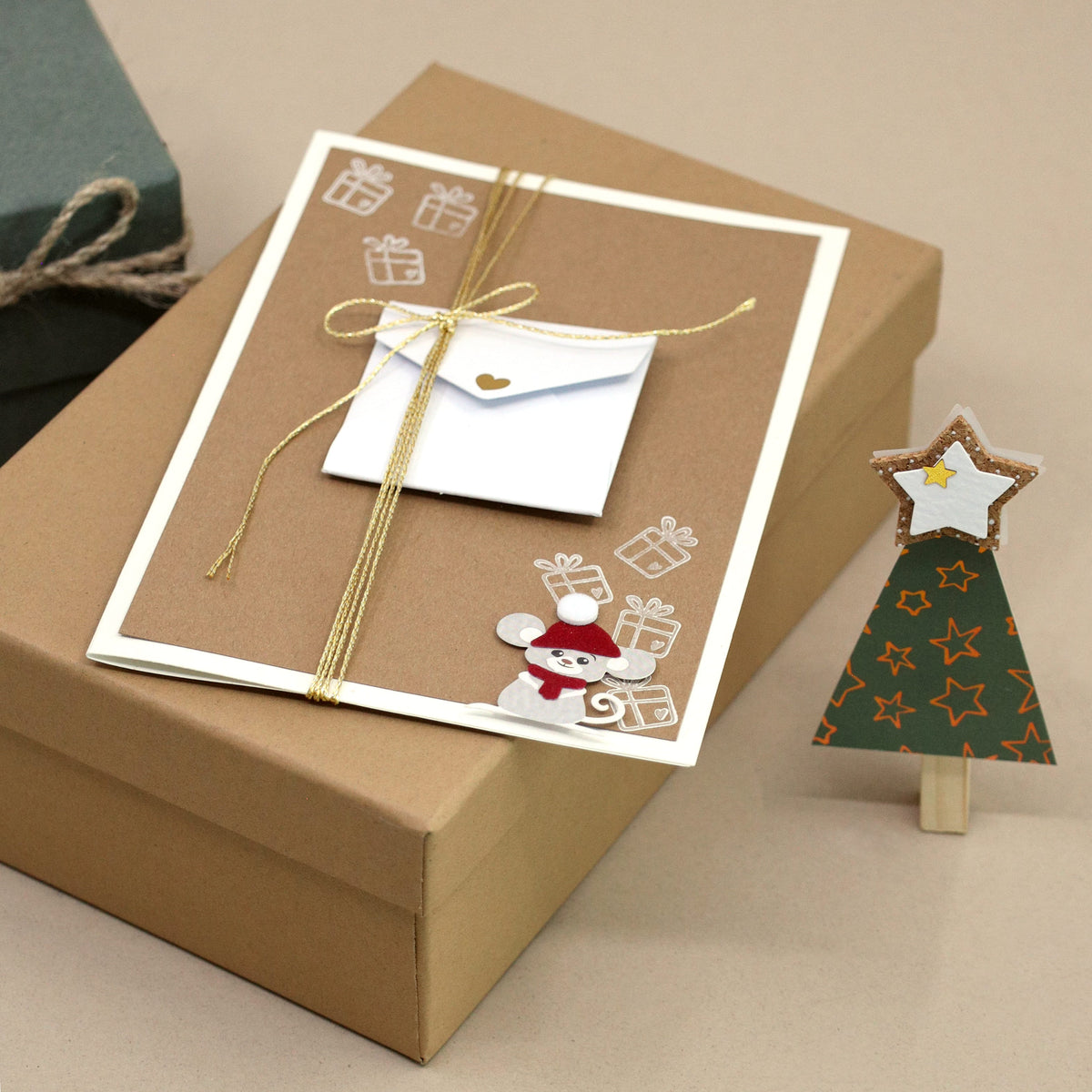 Une carte de Noël ornée de motifs représentant des souris et des cadeaux, accompagnée d'une enveloppe miniature, repose sur un coffret cadeau marron : une touche de choix pour les amateurs d'emballages cadeaux. À côté se trouve le « Stempel Geschenk klein » d'Artoz Papier AG, avec son petit sapin de Noël en papier vert surmonté d'une étoile : une scène charmante qui inspirera vos créations manuelles pour les fêtes.