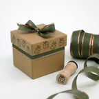 Une boîte-cadeau marron ornée d'un nœud vert et décorée de motifs de cadeaux estampillés à l'aide du tampon « Petit cadeau » de la société Artoz Papier AG. À côté, posés sur un support blanc, se trouvent un tampon en bois – idéal pour les travaux manuels – et un rouleau de ruban assorti.