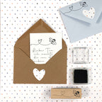 Ein brauner Umschlag mit einem Herzausschnitt enthält eine handgeschriebene deutsche Notiz. Daneben befinden sich ein blauer Umschlag, Bastelarbeiten, ein klarer Stempelblock, ein Stempelkissen und der Artoz Papier AG Stempel Biene - ideal für kreative Einladungskarten.