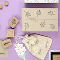 Geschenkverpackungen für Bastelarbeiten auf lavendelfarbenem Untergrund, bestehend aus einer mit Kraftpapier umwickelten Schachtel, die mit dem Stempel Blumen Breeze der Artoz Papier AG bestempelt ist, Holzstempeln mit BRAVO! und Herzen, einem Blumenaufkleber und einem bestempelten Kordelbeutel.