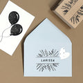 Une enveloppe Einladungskarten bleu pâle avec un art floral et un cœur blanc étiqueté LARISSA repose sur un support brun à côté d'une carte noire et blanche avec deux ballons et le "Stempel Deko für Text" Holzstempel d'Artoz Papier AG.