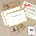Une mise en page festive présentant des articles de papeterie de Noël, notamment des Einladungskarten, le tampon en bois Stempel Engelchen d'Artoz Papier AG, des motifs végétaux, une enveloppe scellée à la cire et une boîte à coussins en forme de champignon - idéal pour Bastelarbeiten.