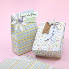 Des sacs et des boîtes à cadeaux aux motifs floraux et à rayures pastel sont disposés sur un fond rose. Chacun d'eux est muni d'une étiquette décorative avec des fleurs, des papillons, des Bastelarbeiten et le tampon Artoz Papier AG Stempel Happy Birthday pour une touche créative.