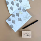 Draufsicht auf Geburtstagskarten mit Ballonmotiven und "Happy Birthday"-Text, daneben ein schwarzer Stift und der Artoz Papier AG Stempel Happy Birthday mit Punkten - ideal für Bastelarbeiten oder um eine persönliche Note hinzuzufügen.