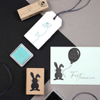 Une présentation à plat des fournitures de Bastelarbeiten, comprenant l'Artoz Papier AG Stempel Osterhase, des tampons à motifs floraux, des tampons encreurs, une étiquette cadeau à fleurs bleues tamponnée, et une carte avec un lapin noir et "Für Jeaninne" - idéale pour les Einladungskarten.