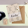 Carte de Pâques avec un lapin dessiné à la main sur du papier brun, accompagnée d'une enveloppe assortie. Une marguerite blanche orne le set, présenté avec le Stempel Hase d'Artoz Papier AG et un tampon encreur noir - idéal pour les travaux manuels ou les invitations. Le mot "Pâques" est inscrit sur la carte.