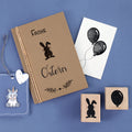 Une carte brune "Frohe Ostern" avec un lapin, une carte postale à deux ballons noirs, un porte-clés licorne et deux pochoirs Artoz Papier AG Stempel Osterhase - parfaits pour Bastelarbeiten ou Einladungskarten - sont placés sur un fond bleu.