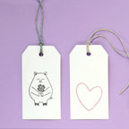 Deux étiquettes cadeaux blanches d'Artoz Papier AG sur fond violet. L'étiquette de gauche représente un cochon tenant des fleurs pour Bastelarbeiten, tandis que l'étiquette de droite reprend le motif Stempel Herz asymmetrisch en rouge. Les deux étiquettes sont munies de boucles de ficelle.